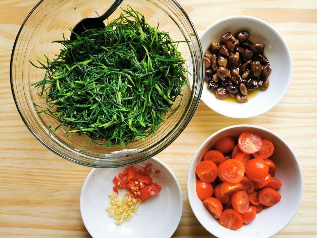 halved cherry tomatoes in white bowl. Blanched agretti in white bowl. chopped garlic and peperoncino in white bowl and pitted olives in white bowl.