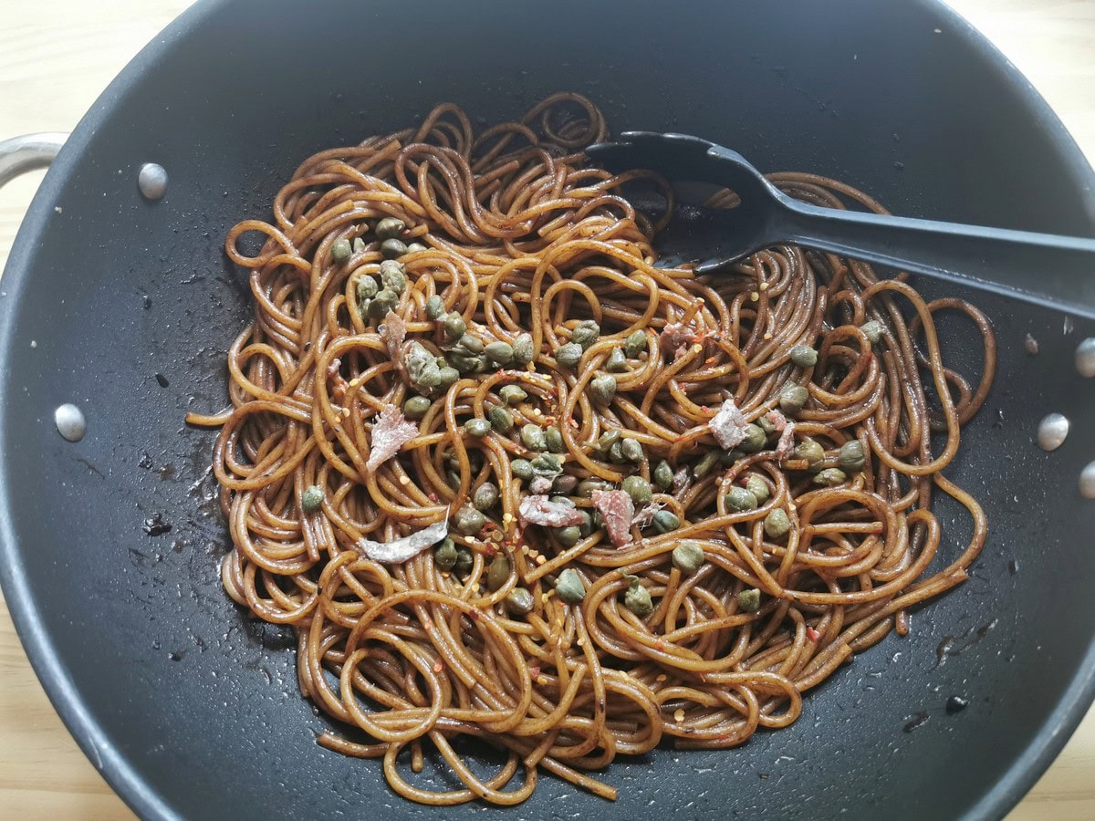Capers, anchovy pieces and peperoncino flakes added to black garlic spaghetti in skillet.