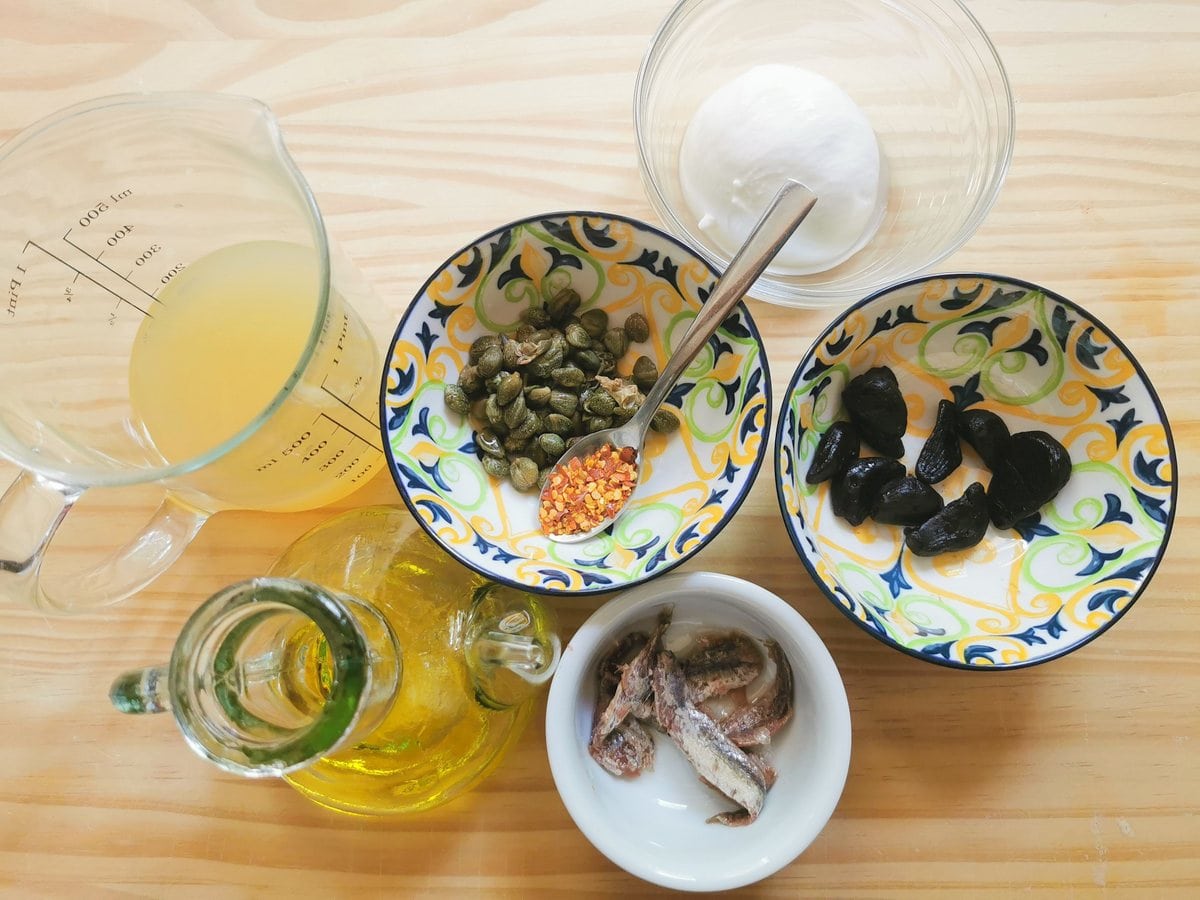 Ingredients for black garlic sauce on wood work top; black garlic cloves, salted anchovies, olive oil, chicken broth, salted capers and peperoncino flakes.