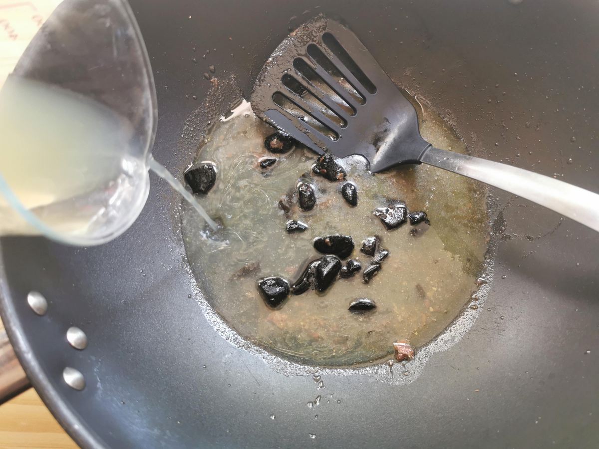 Chicken broth being poured into large skillet with dissolved anchovies and black garlic.