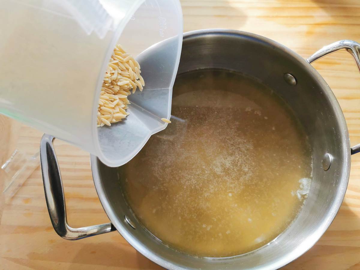 Dried orzo pasta being poured into hot beef broth in cooking pot.