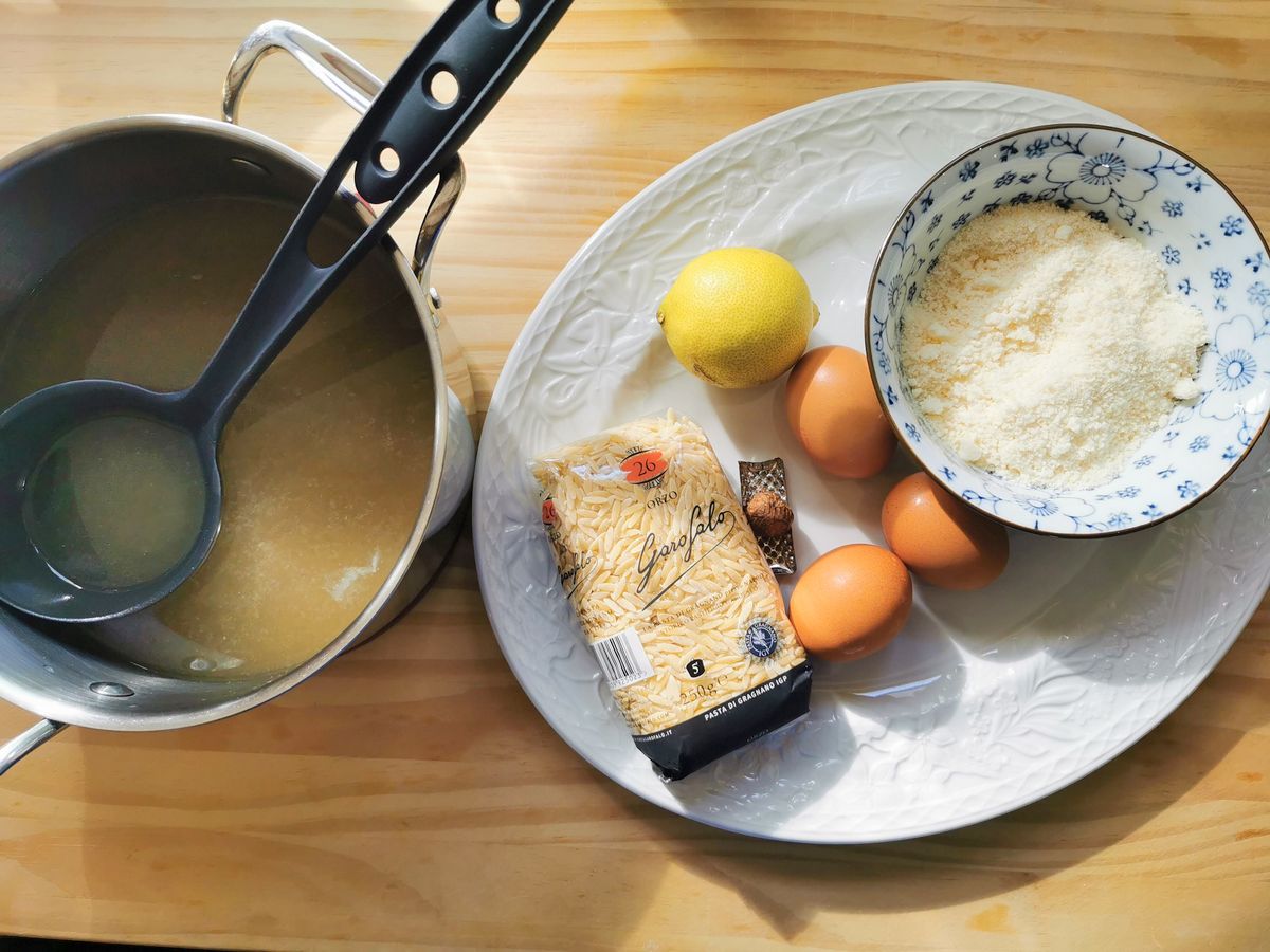 Ingredients for stracciatella soup: orzo pasta, parmigiano cheese, eggs, nutmeg and a lemon on a white plate and beef broth in a pan.