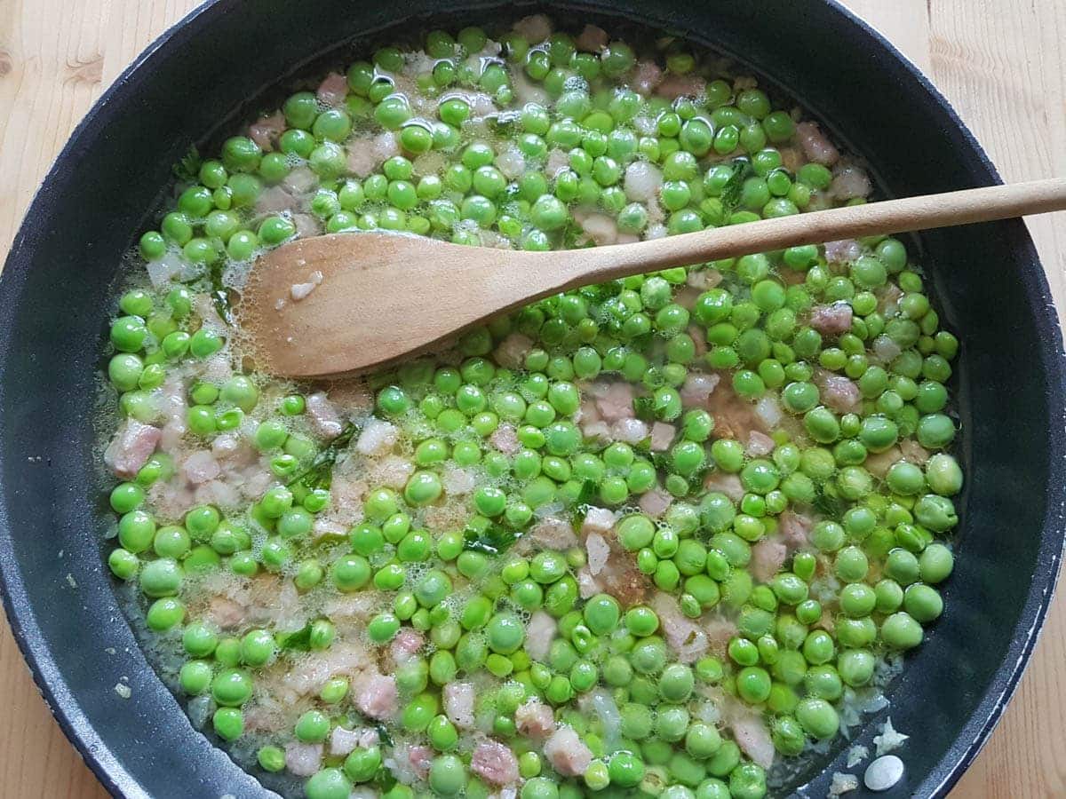 Fresh peas, pancetta and onion cooking in broth in frying pan