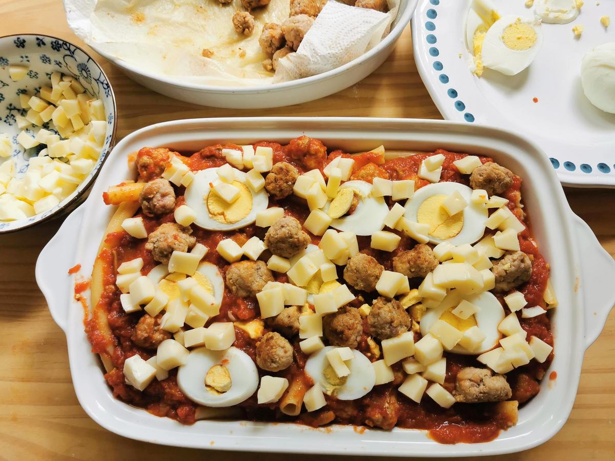 Ziti alla lucana in oven dish with cheese cubes, meatballs and hardboile eggs on top.