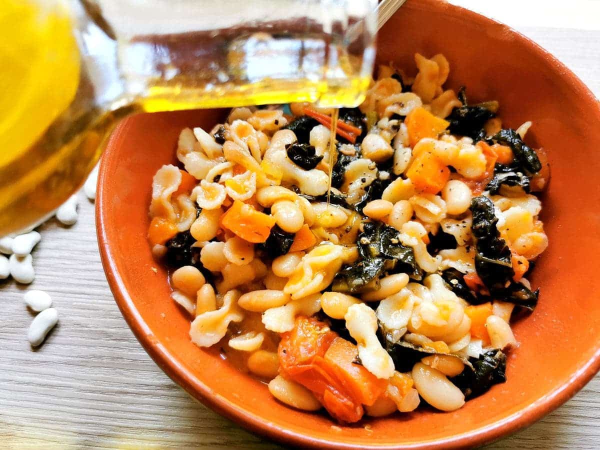 Tuscan kale, white bean and pasta soup in terracotta bowl with olive oil being added