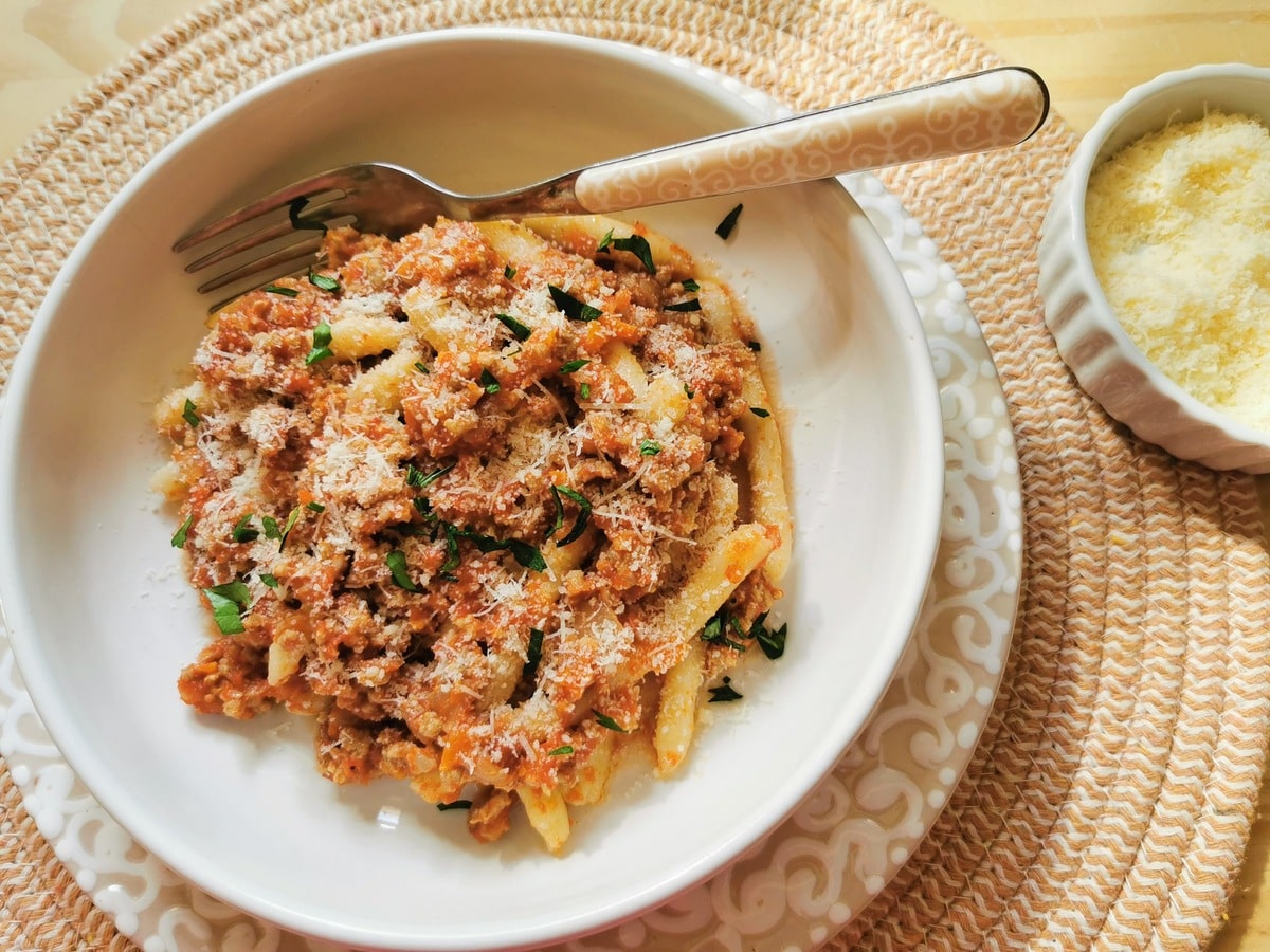 Veal ragu with homemade maccheroni sprinkled with grated Parmigiano and fresh parsley in white bowl.