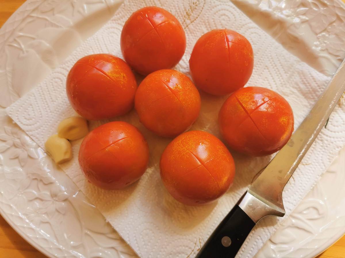 Fresh tomatoes with an 'x' cut into the peel plus, peeled garlic cloves on white plate.