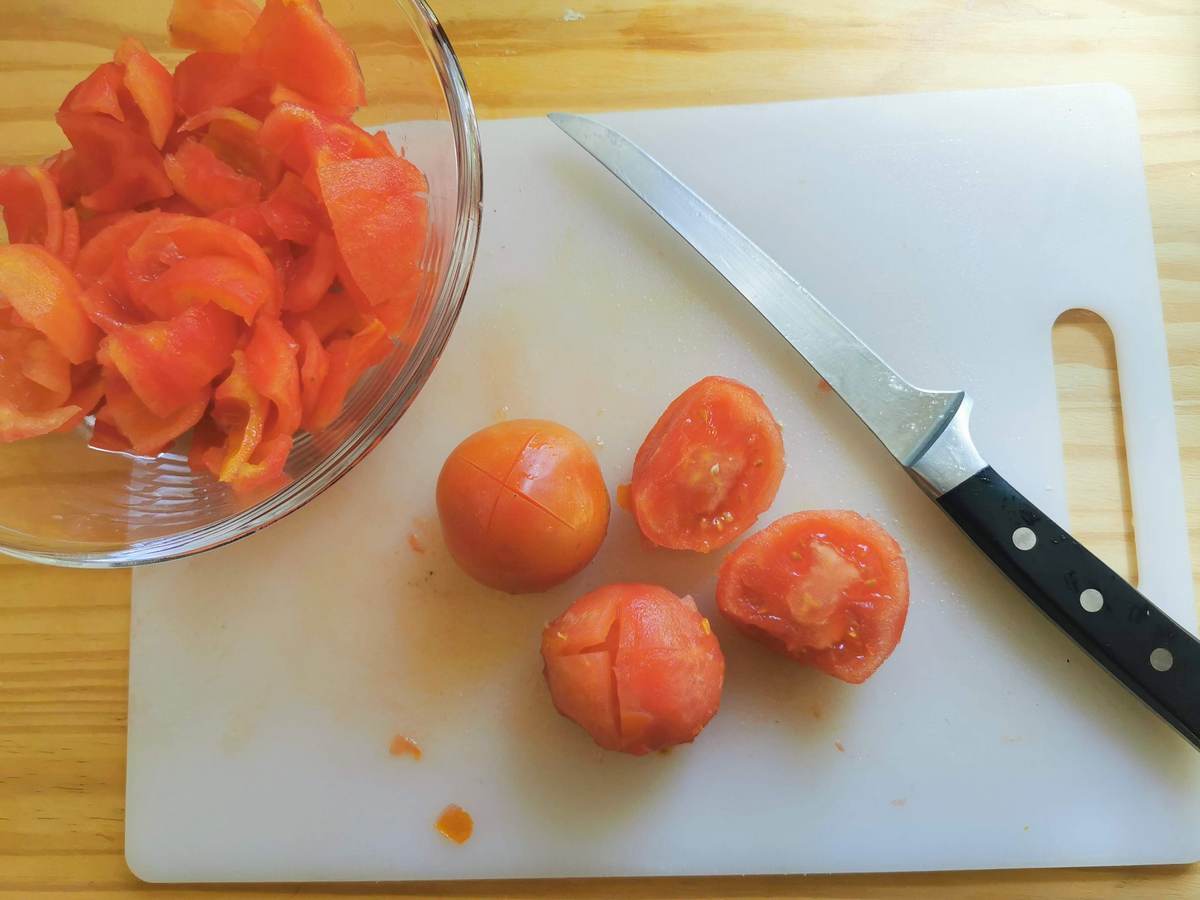 Peeled tomatoes on white chopping board and peeled and deseeded tomato pieces in glass bowl.