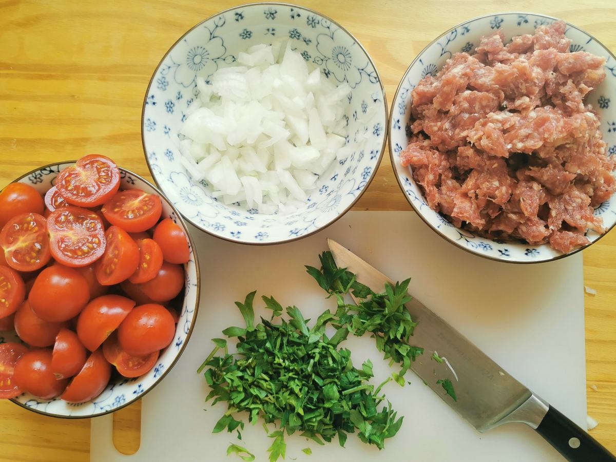 Prepared ingredients for luganega sausage ragu: halved cherry tomatoes, crumbled sausage and finely chopped onion in small bowls.