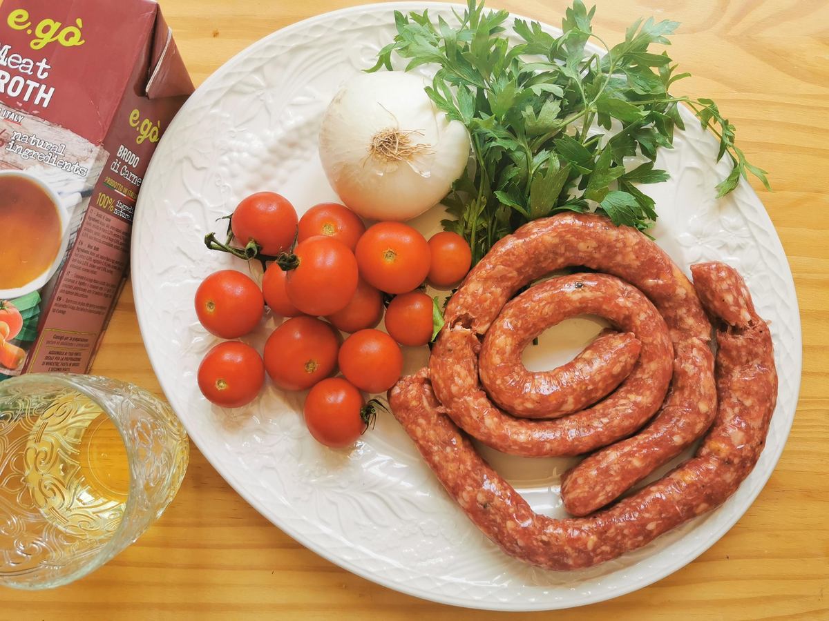 Ingredients for luganega sausage ragu on white plate: cherry tomatoes, fresh parsley, white onion and a luganega sausage spiral.