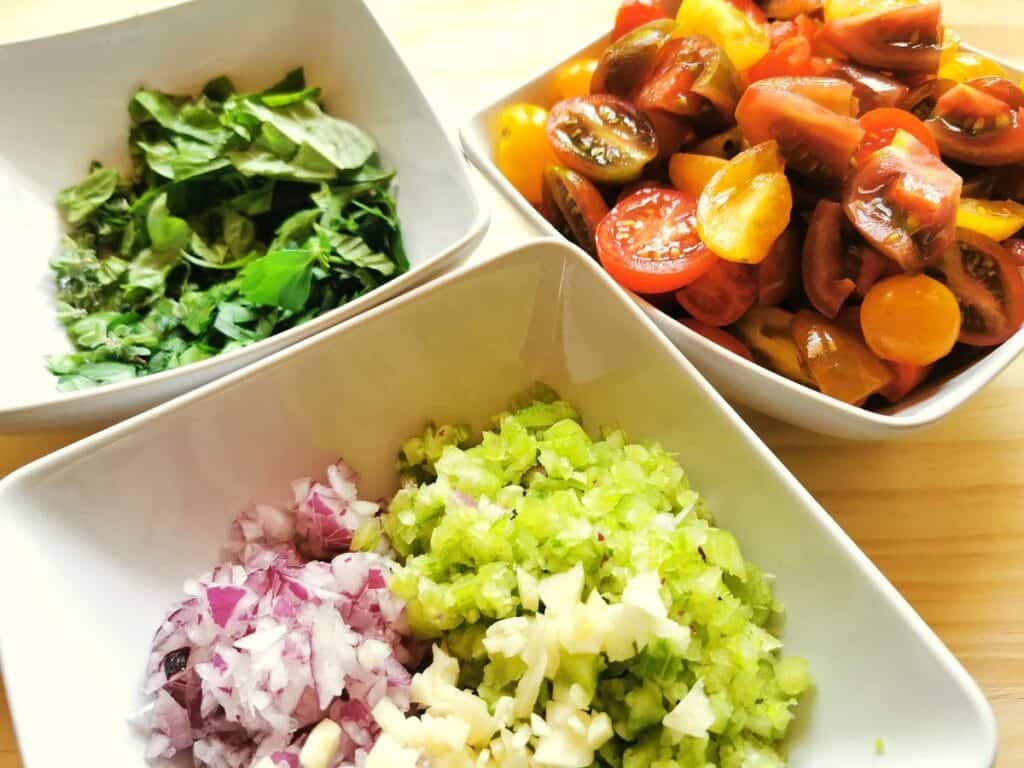 chopped onions, celery and garlic in white bowl. Chopped tomatoes in white bowl. Chopped parsley in white bowl