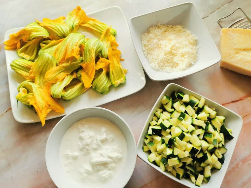 cut zucchini flowers, grated Parmigiano, stracciatella cheese and cubed zucchini in white bowls