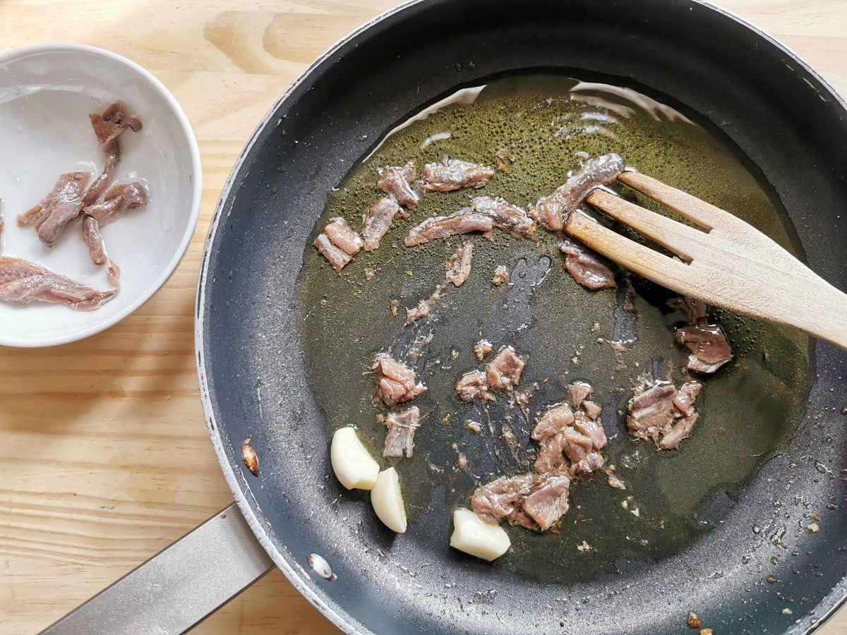 Pieces of anchovies and peeled garlic cloves in skillet with olive oil.