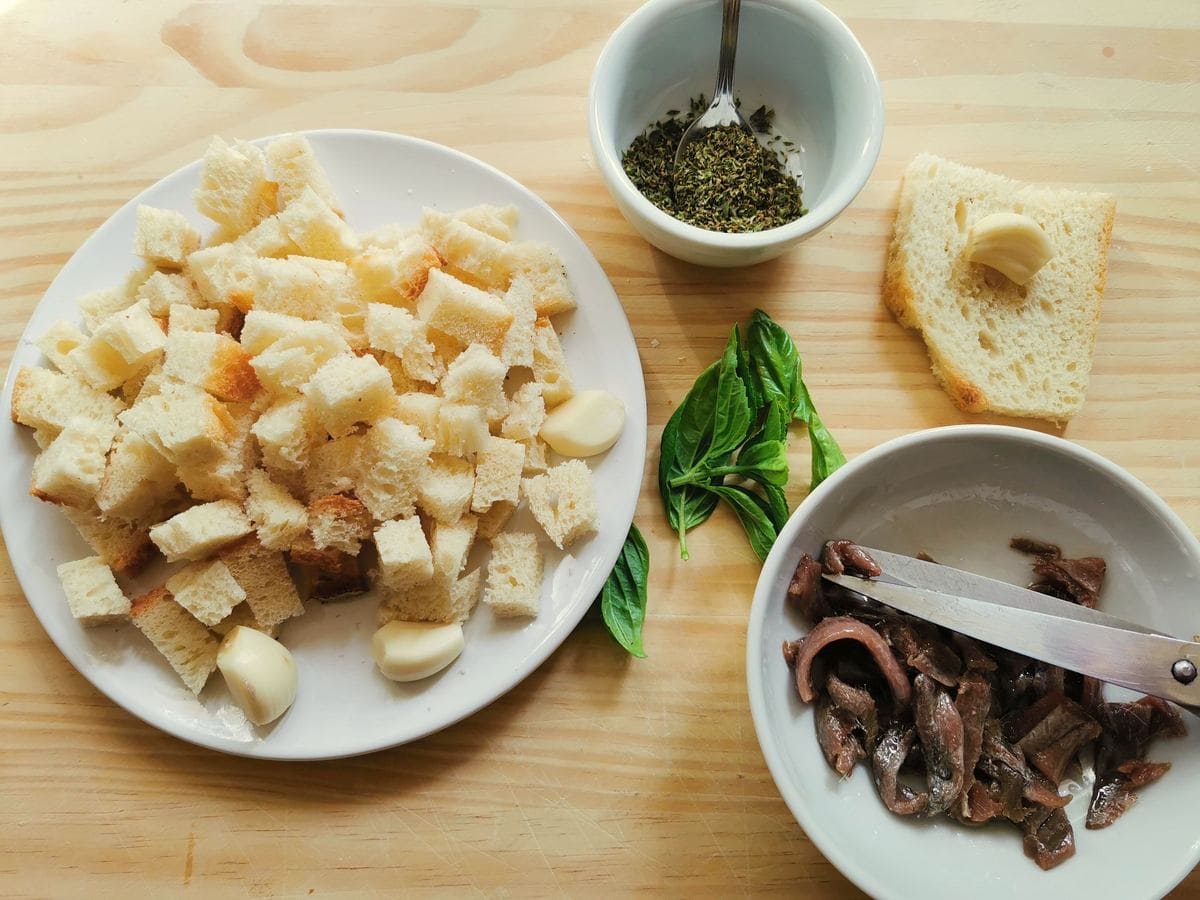 Pieces of anchovies in white bowl, Bread cubes on white plate and dried oregano in small white bowl.