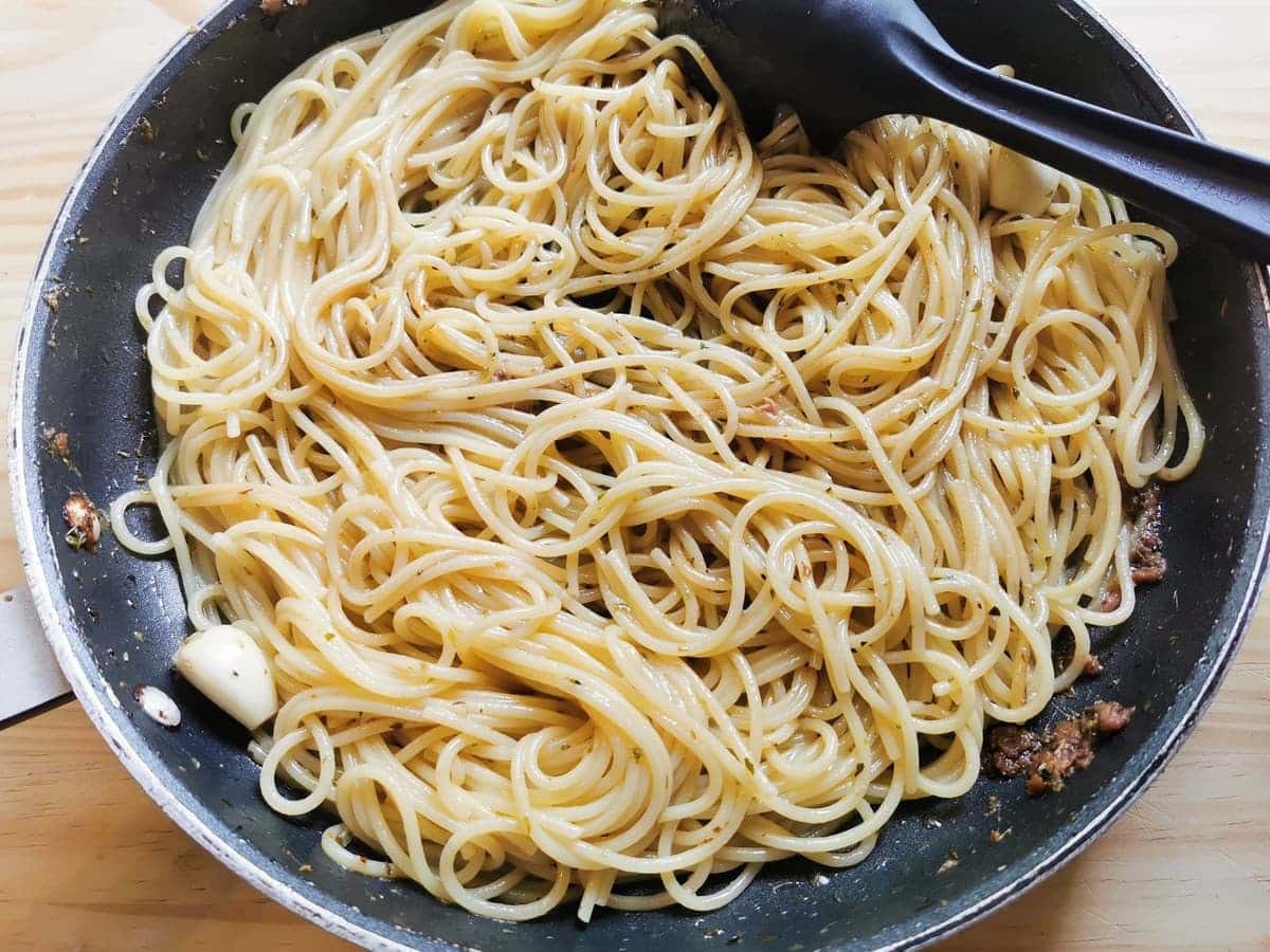 Cooked spaghetti mixed with anchovies, garlic and oregano in skillet.