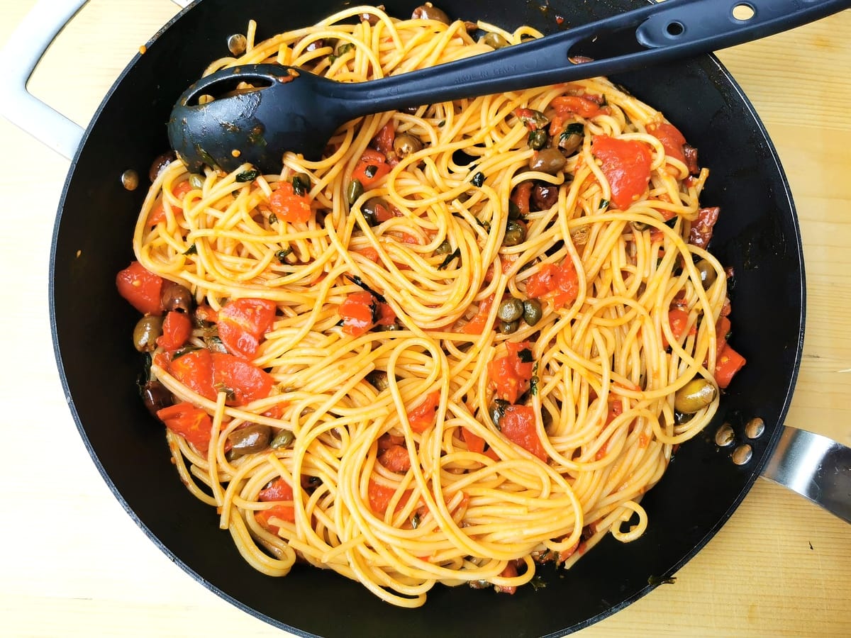 Cooked spaghetti in pan with puttanesca sauce ingredients.