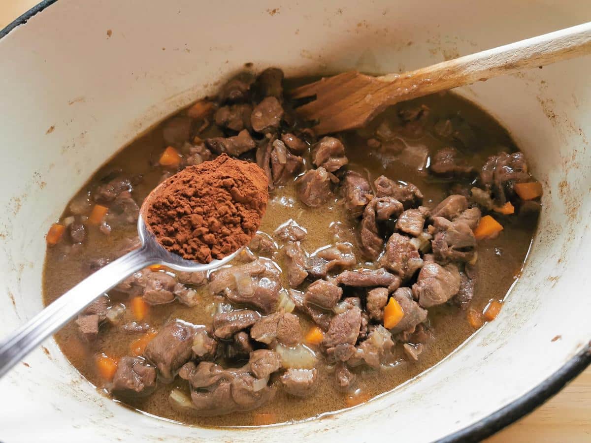 A tablespoon of cocoa powder being added to hare ragu in Dutch oven.