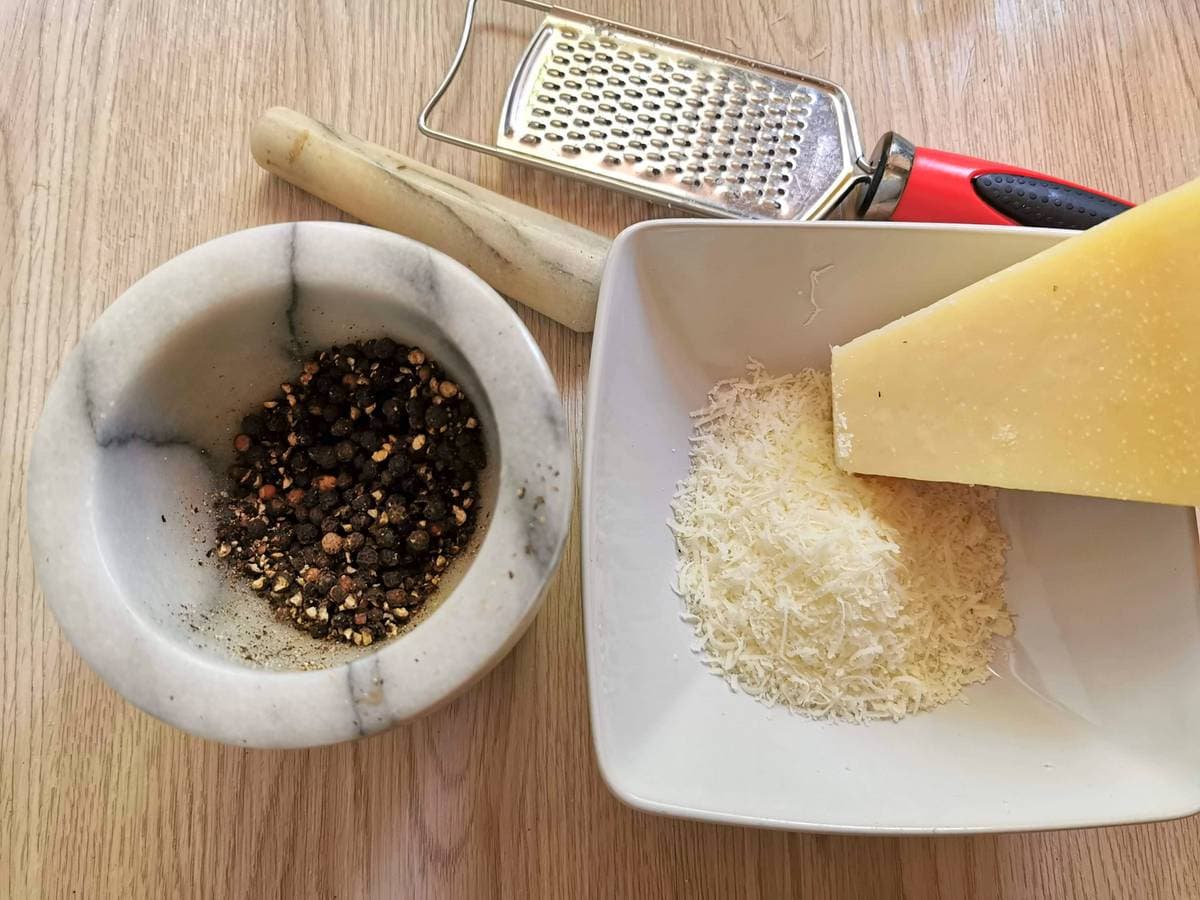 Crushed pepper corns in a marble mortar and grated Pecorino Romano in a white bowl.