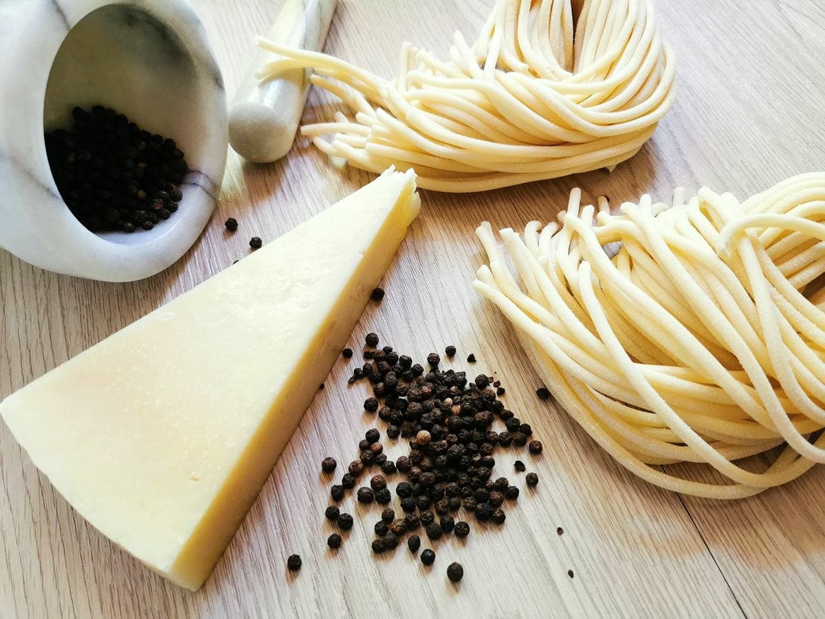 ingredients for cacio e pepe; fresh thick spaghetti, black pepper corns and Pecorino Romano cheese on wood worktop.