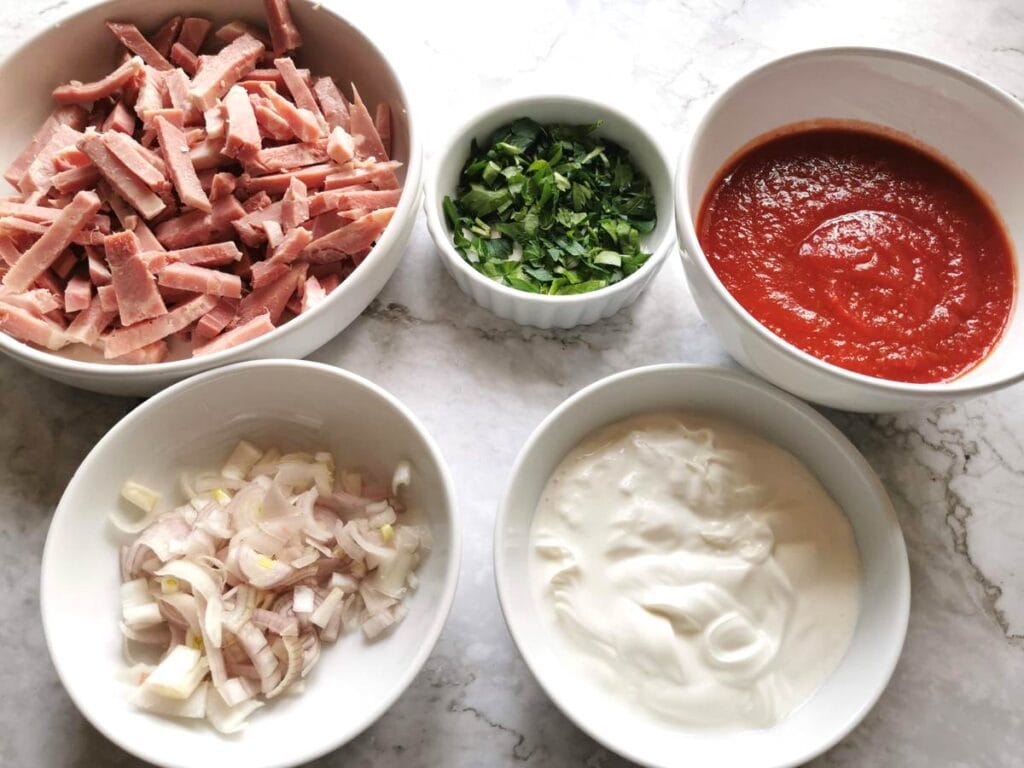 Sliced shallots, chopped ham, chopped parsley, heavy cream and tomato passata in separate white bowls.