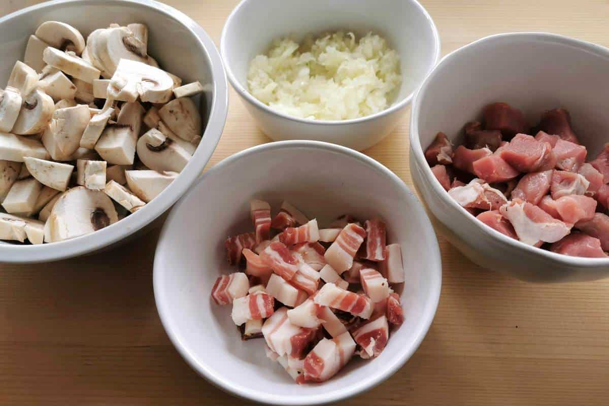 Cut mushrooms, onions, pancetta and pork fillet in white bowls.