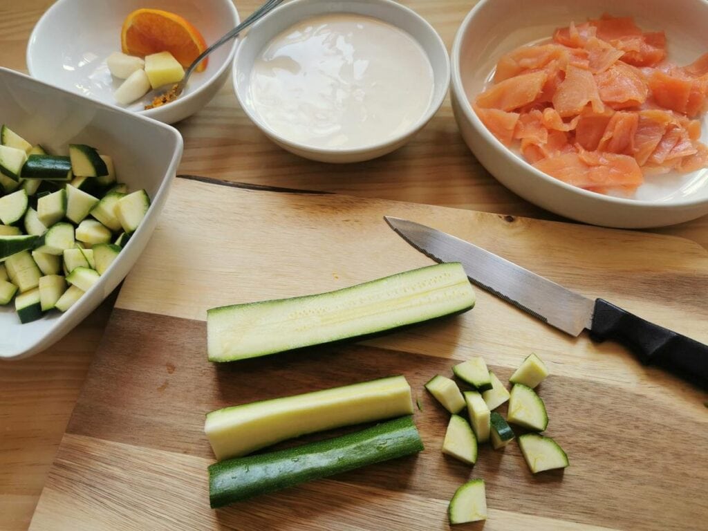 cut zucchini on bread board and in white bowl, cut salmon in white bowl and cream in white bowl