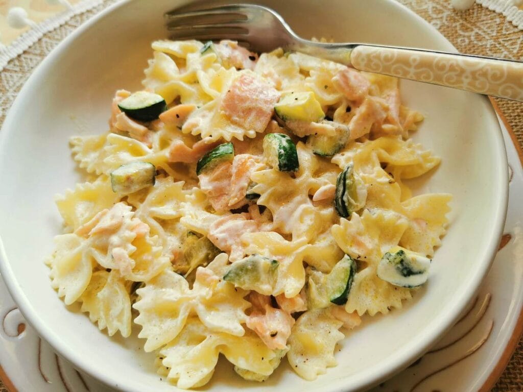 A bowl of bow-tie pasta mixed with creamy sauce, zucchini slices, and chunks of salmon, served with a fork. The dish is presented on a lace tablecloth.