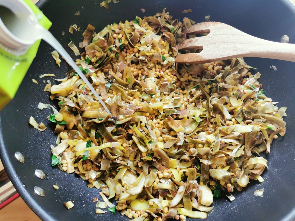 Vegetable broth being poured into the pasta and artchokes in deep frying pan.