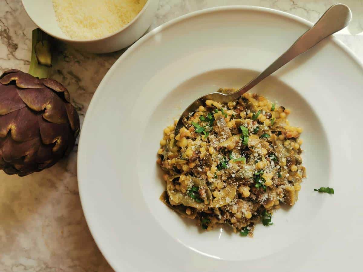 Fregole pasta with artichokes in a white bowl with fresh parsley and grated Pecorino sprinkled on top.