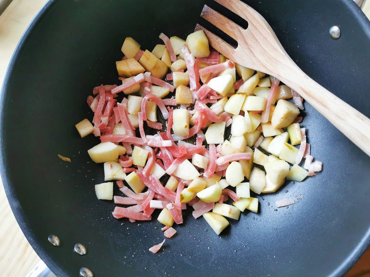 Small strips of speck in deep frying pan with pieces of pear.