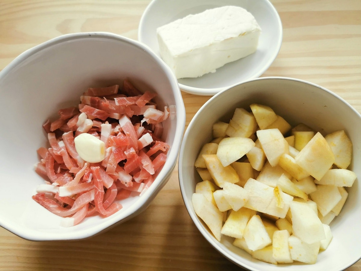 Cubes of peeled pears in white bowl, small pieces of speck and peeled garlic clove in white bowl. Piece of taleggio in white bowl.