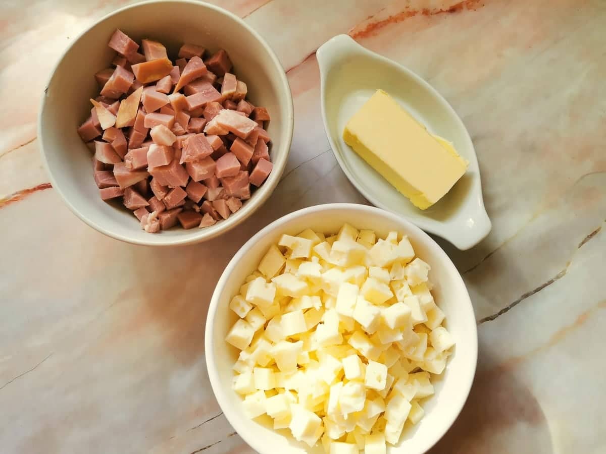 Cubes of cheese in white bowl, cubes of ham in white bowl and piece of butter in white bowl.