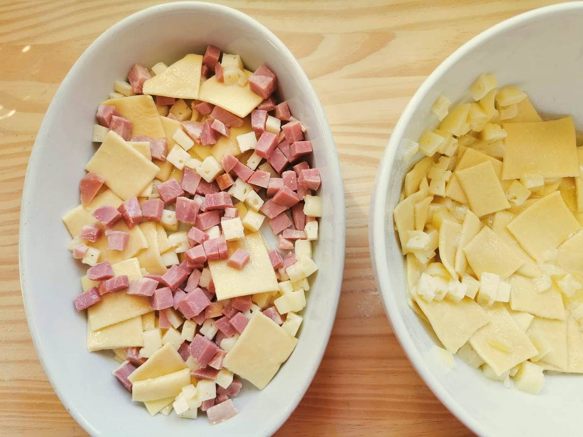 White oval oven dish with pasta squares and ham and cheese cubes next to white bowl with fresh pasta squares and cheese cubes.