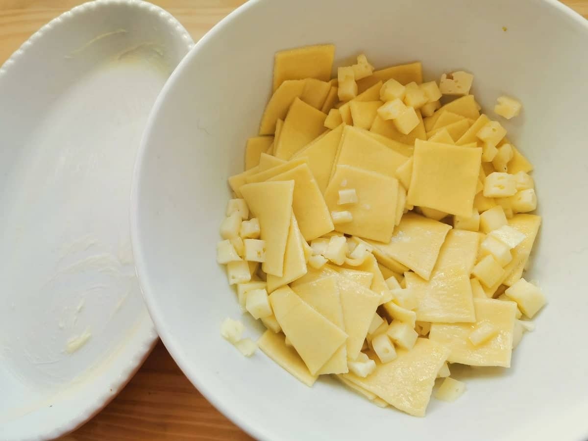 Partly cooked pasta squares in white bowl with melted butter and cubes of Asiago cheese.