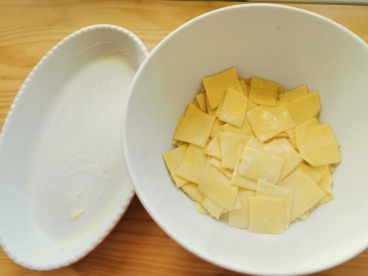 Partly cooked pasta squares in a white bowl and oval oven dish greased with butter.