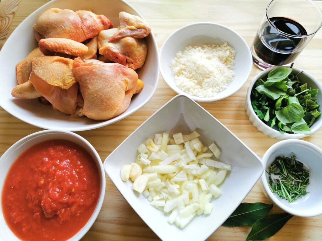 chopped onions and garlic in white bowl. Chopped herbs in white bowl and quartered chicken in white bowls. Plus tomato pulp in white bowl