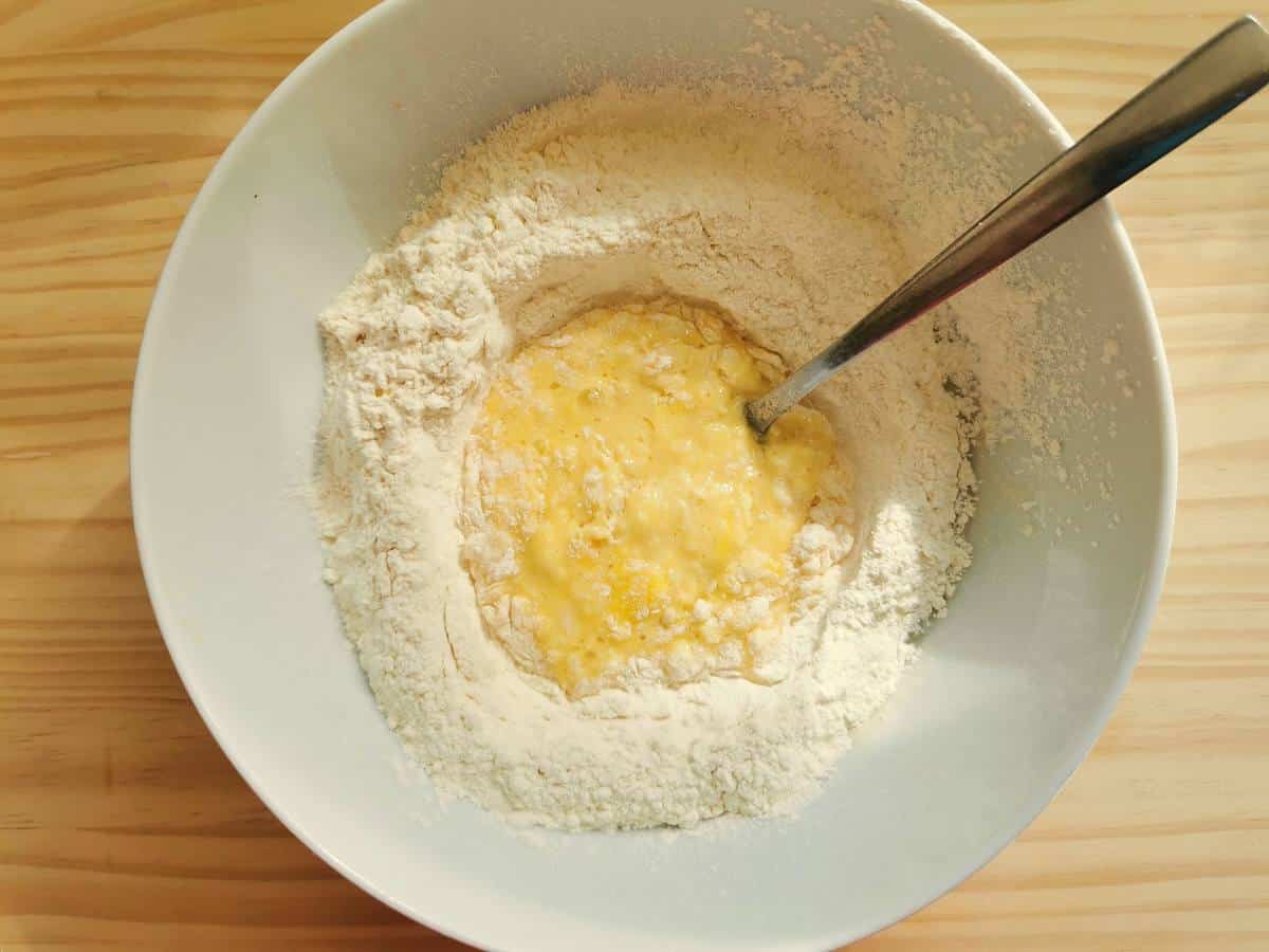 Eggs beaten with a fork in the middle of white flour in a hite bowl.