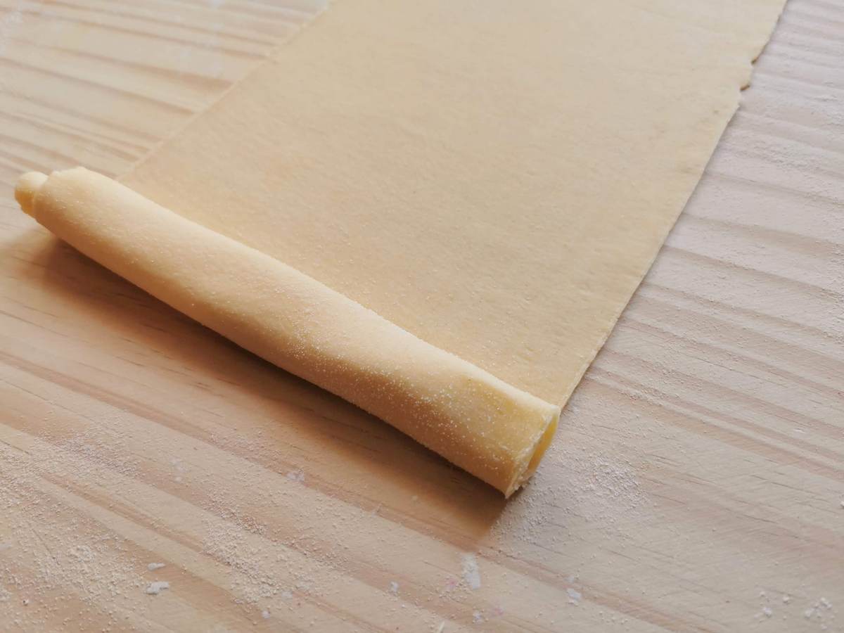 Tagliatelle pasta dough sheet being rolled up on wooden board.