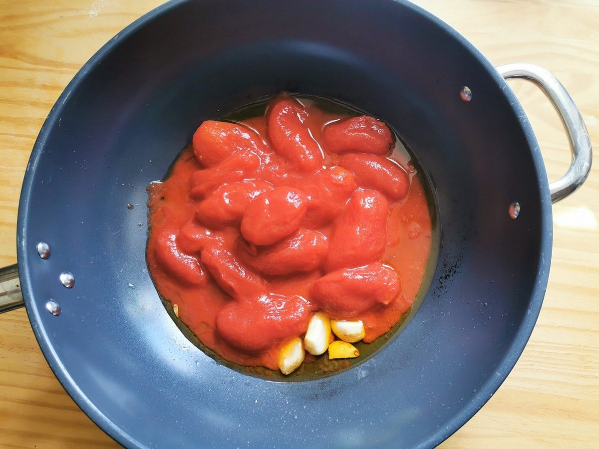 Canned and peeled datterini tomatoes in deep skillet with 3 peeled garlic cloves.