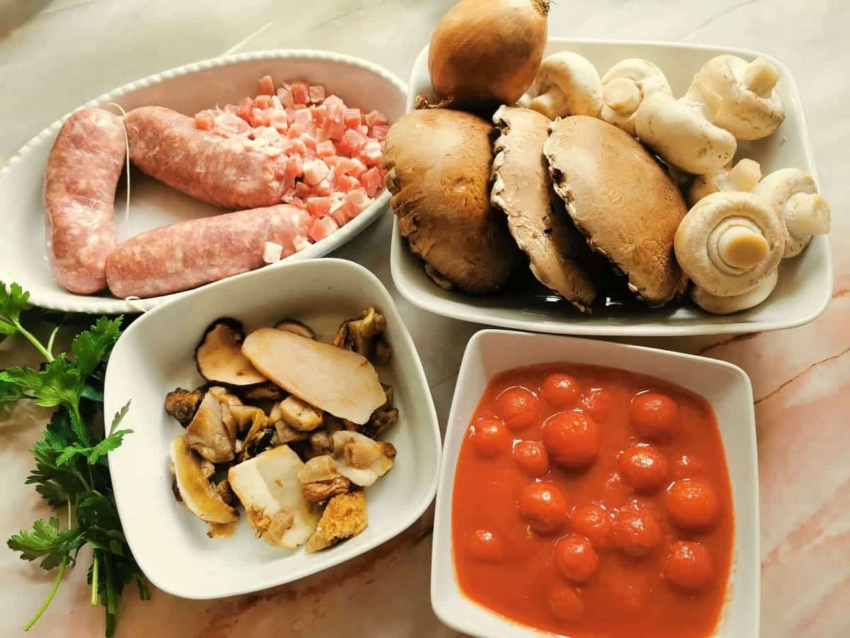 Ingredients for pasta alla boscaiola with sausage and mushrooms in white bowls on marble worktop.