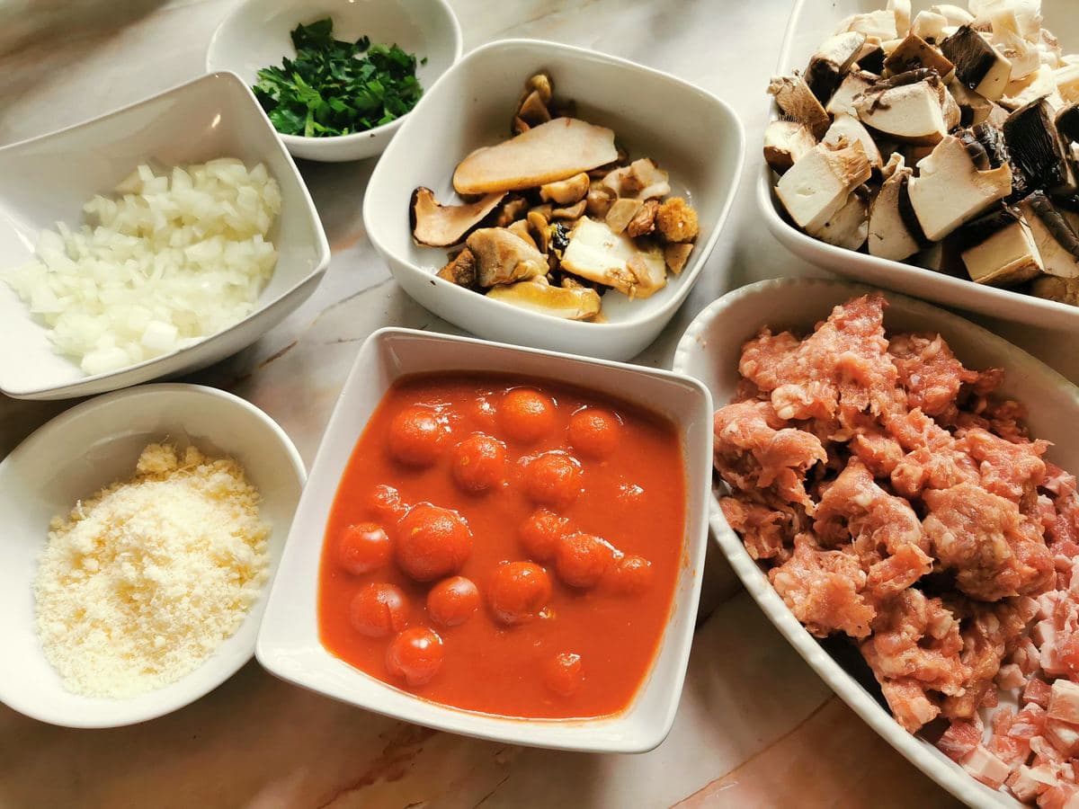 Broken up sausage meat and pancetta in white bowl. Chopped onion in white bowl. Cut mushrooms in 2 white bowls. Canned tomatoes in white bowl.