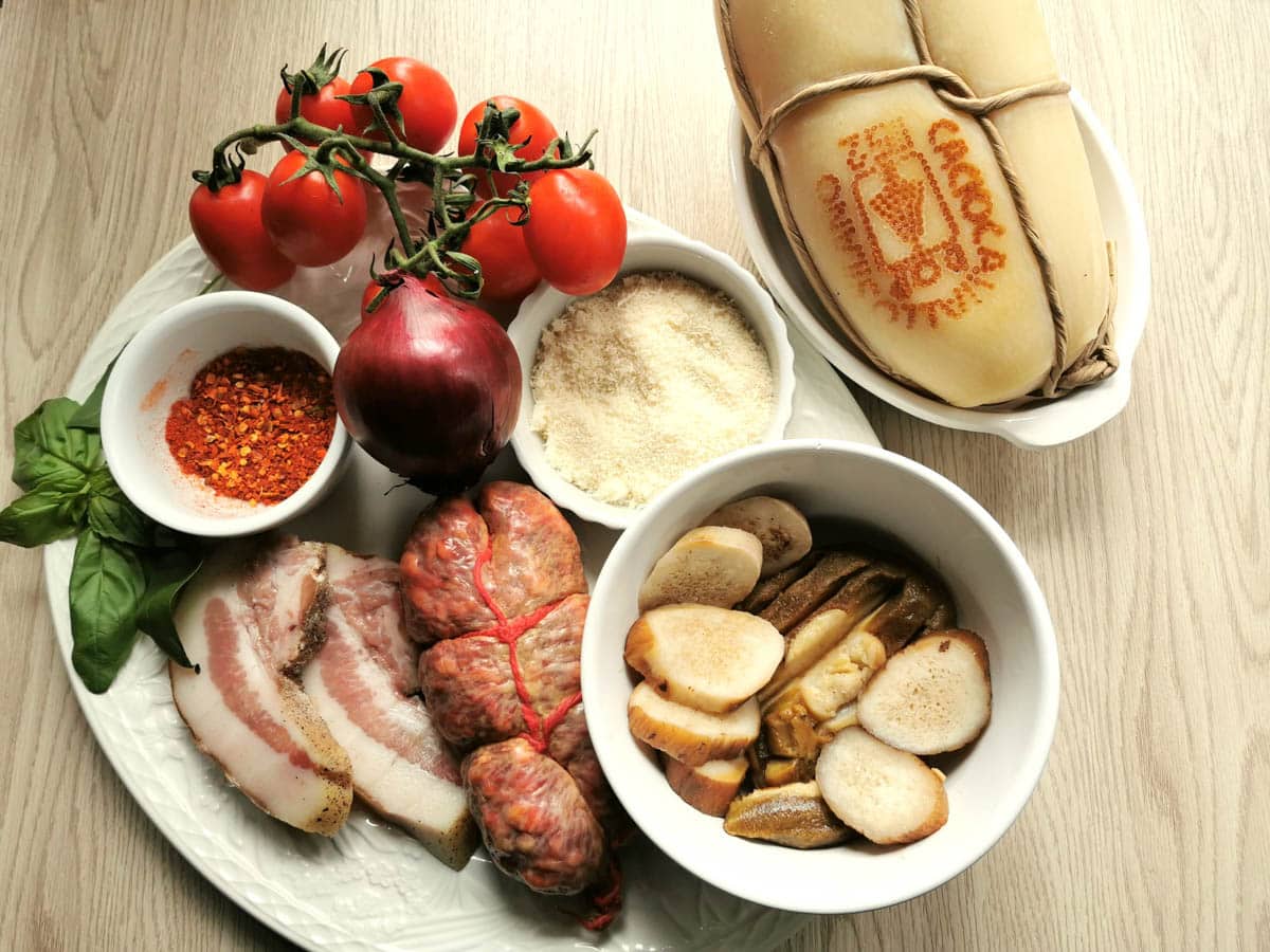 Unprepared ingredients on a kitchen counter: fresh tomatoes, guanciale, soppressata, caciocavallo, red onion, porcini, pecorino cheese, basil leaves, peperoncino flakes.