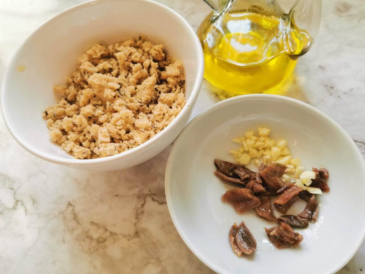 Breadcrumbs in white bowl. Chopped garlic and anchovies in white bowl and Glass jug with olive oil.