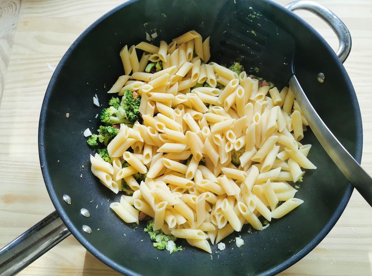 Cooked penne pasta with broccoli and pancetta in frying pan.