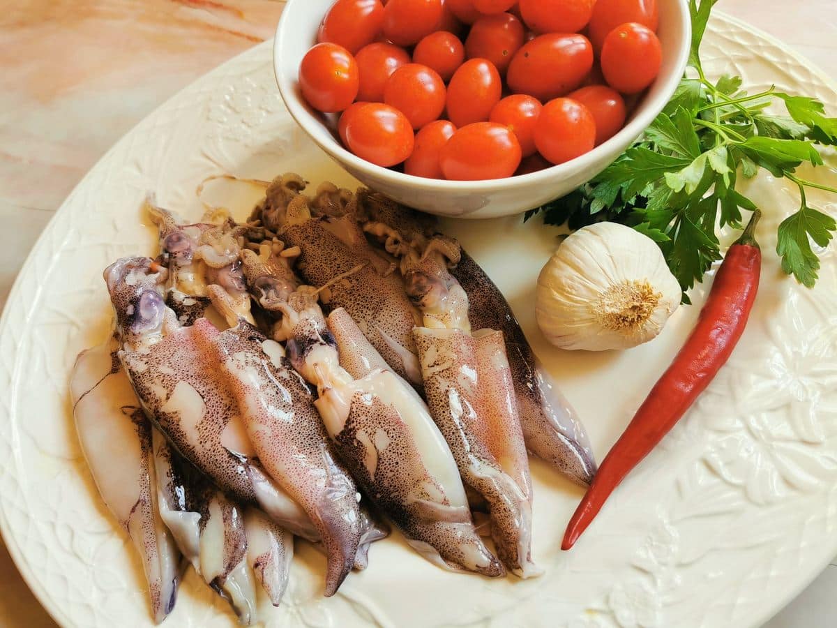 Fresh calamari, datterini tomatoes, garlic, red chilli pepper and fresh parsley on white plate.