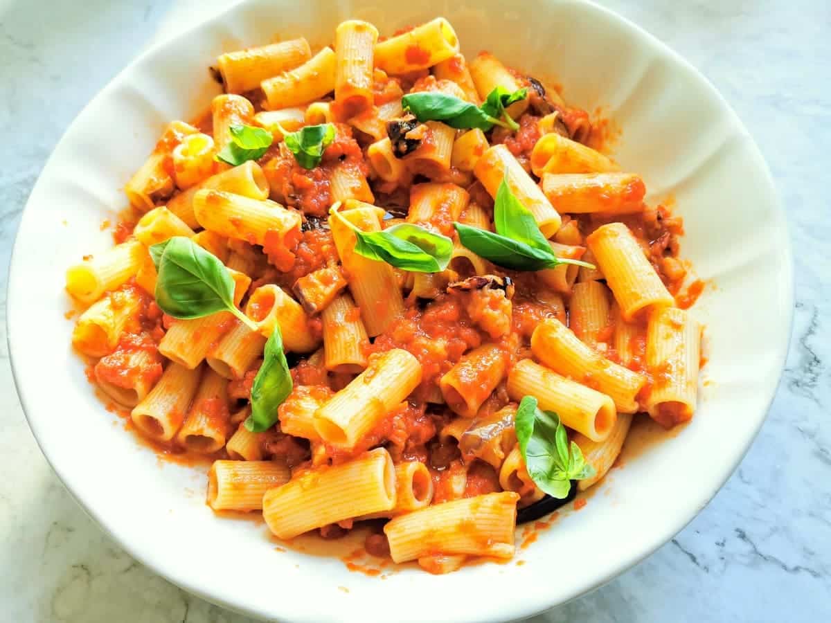 Cooked pasta in a bowl mixed with tomato and eggplant sauce with basil leaves on top.