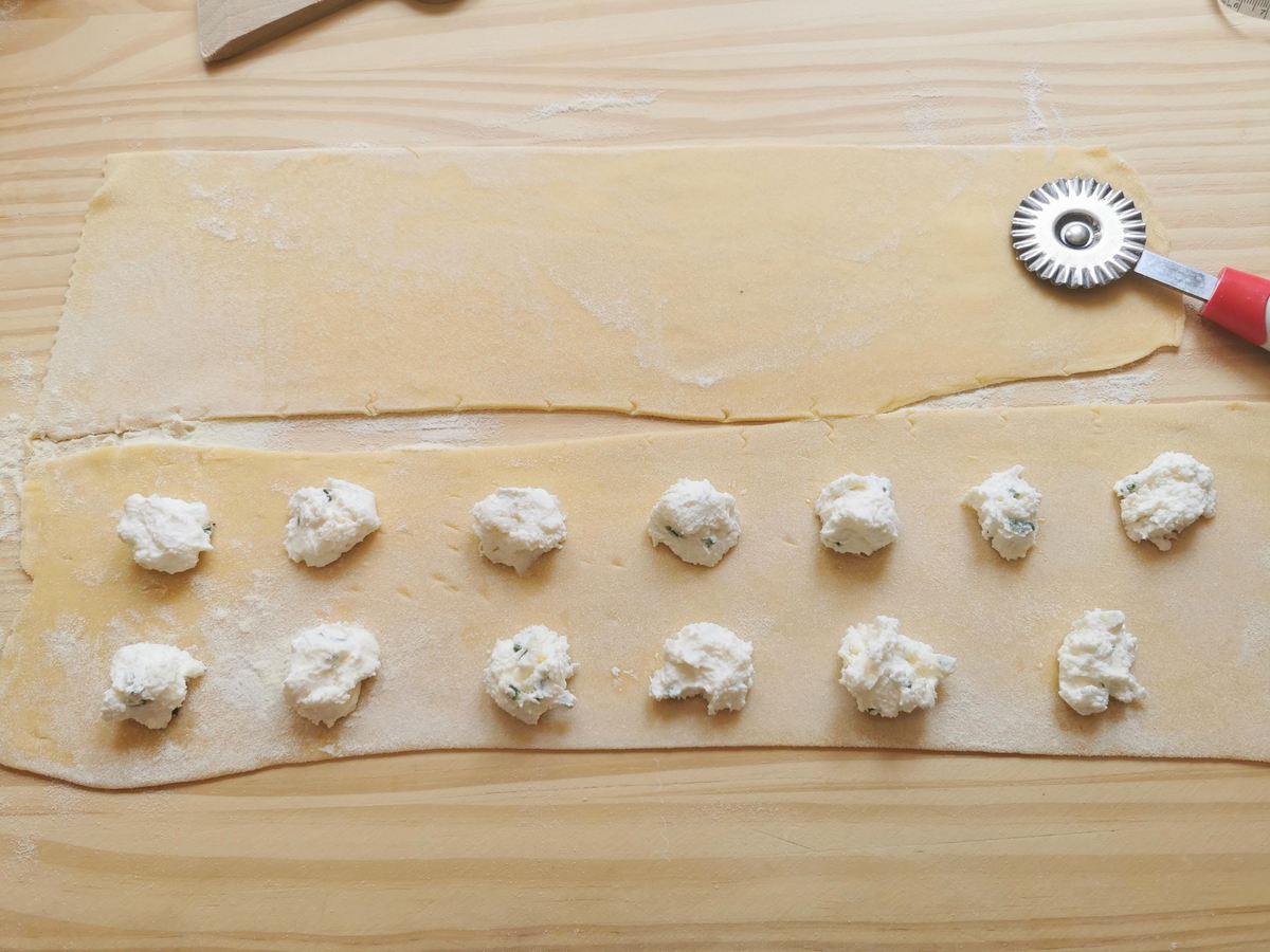Two sheets of rolled out pasta dough on wood work surface. One sheet has teaspoons of filling on it.