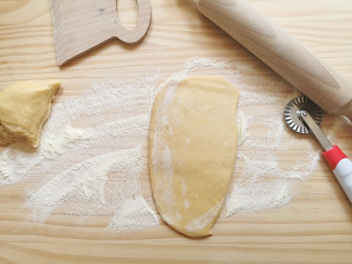 Flattened piece of pasta dough on flour dusted work surface with a rolling pin and ravioli cutter.