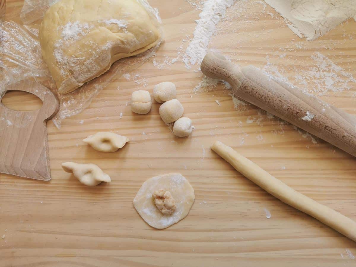 Stages for making scarpinocc on a wood work surface. Dough rope, gnocchi size pieces of dough, circle of dough with filling and ready scarpinocc.