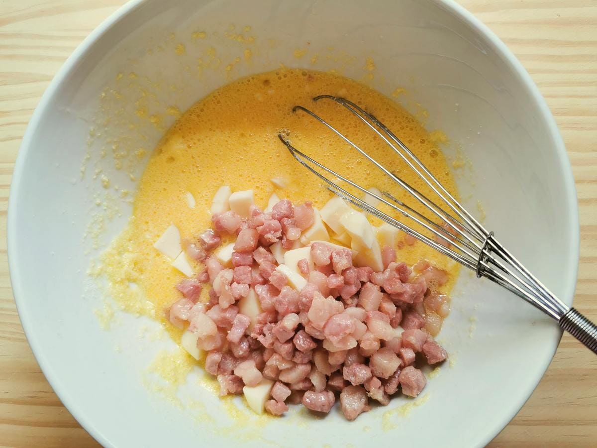 Cut scamorza cheese and cubed pancetta in white bowl with eggs and grated Parmigiano cheese.